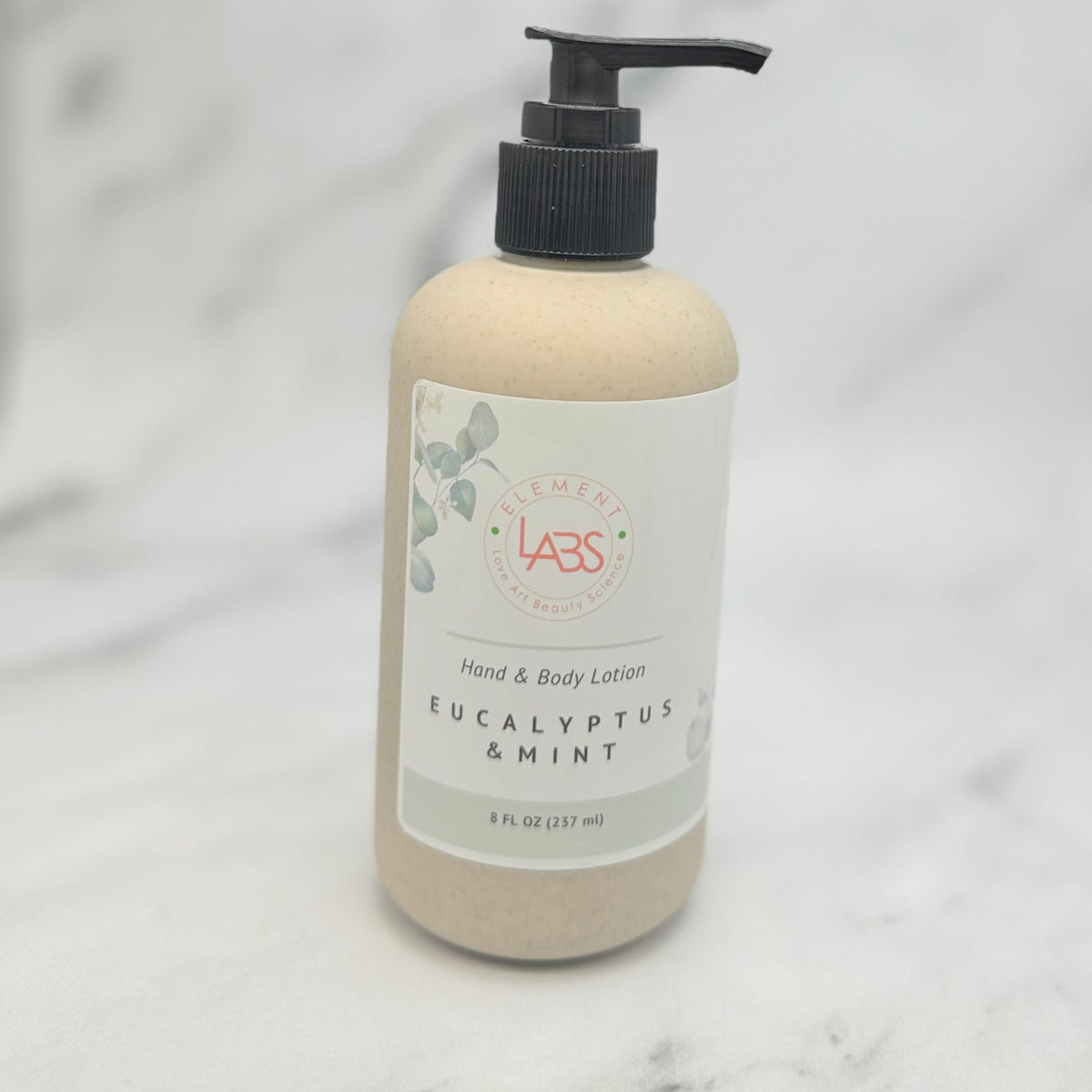 A bottle of Element LABS Hand & Body Lotion in Eucalyptus & Mint, featuring a beige textured body, a white label with green and red details, and a black pump dispenser, set against a light gray marble background.