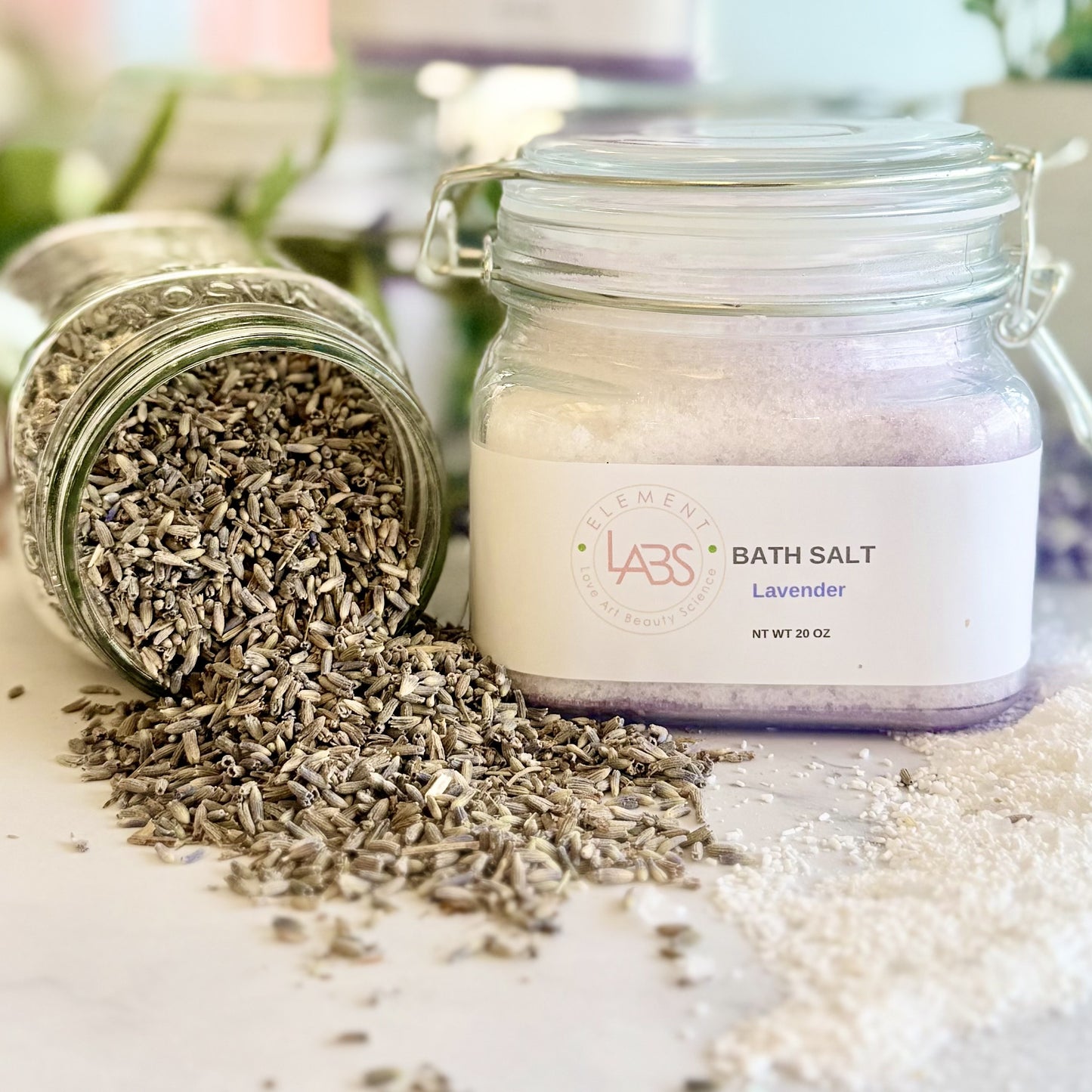 A glass jar labeled Element LABS BATH SALT Lavender' stands on a light surface with loose dried lavender seeds spilled from another tipped jar beside it.