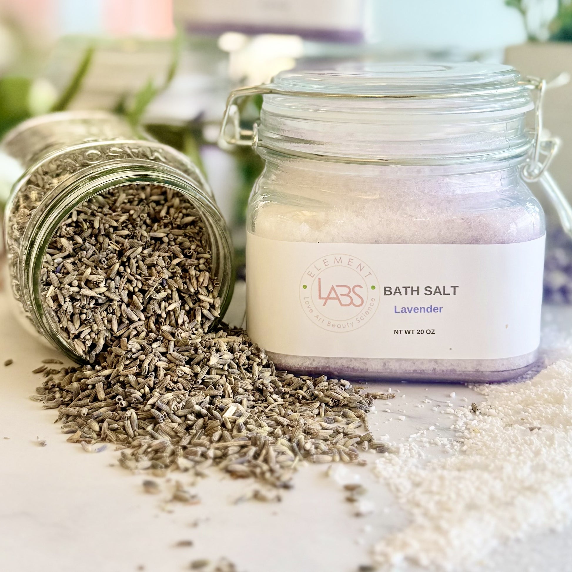 A glass jar labeled Element LABS BATH SALT Lavender' stands on a light surface with loose dried lavender seeds spilled from another tipped jar beside it.