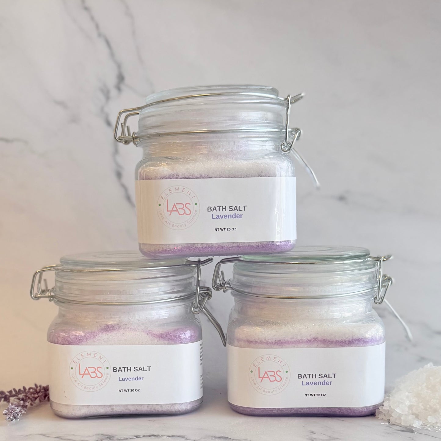 Three glass jar bath salts with lavender labels, stacked in a pyramid arrangement on a white marble surface with a marble background.