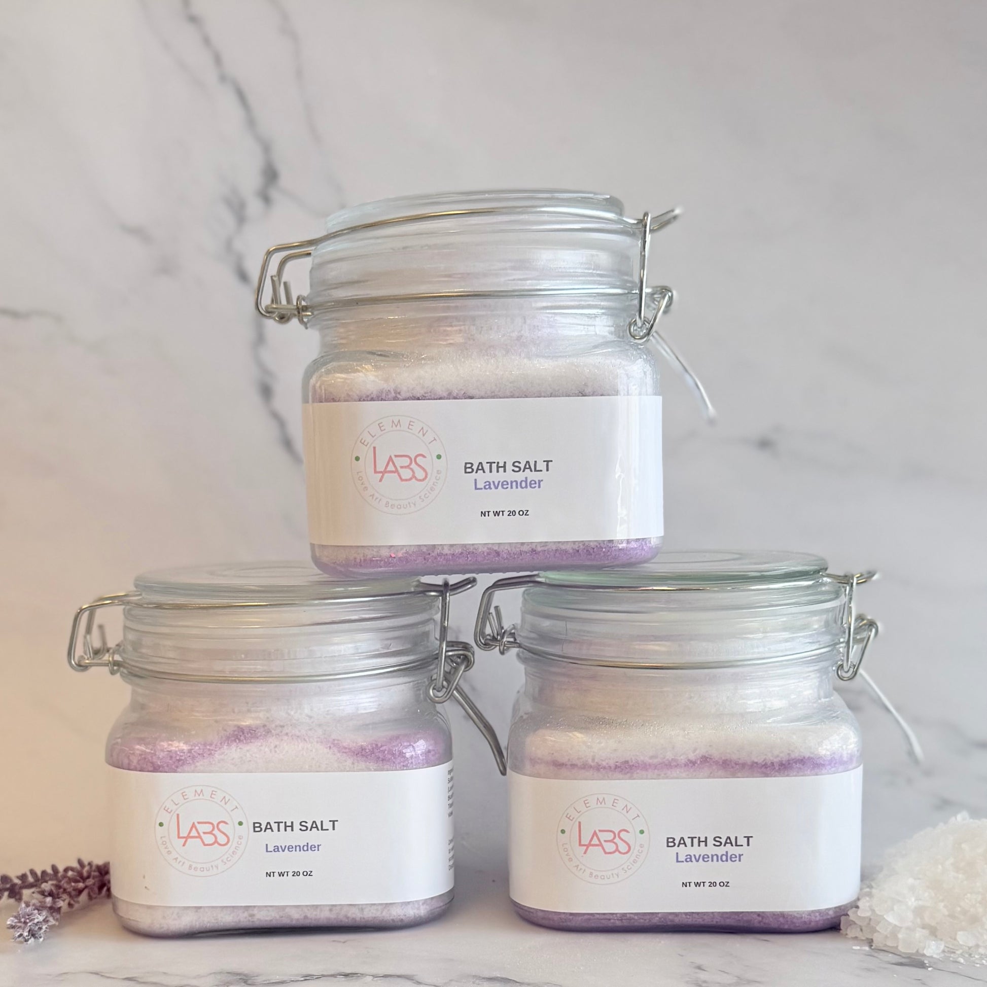 Three glass jar bath salts with lavender labels, stacked in a pyramid arrangement on a white marble surface with a marble background.