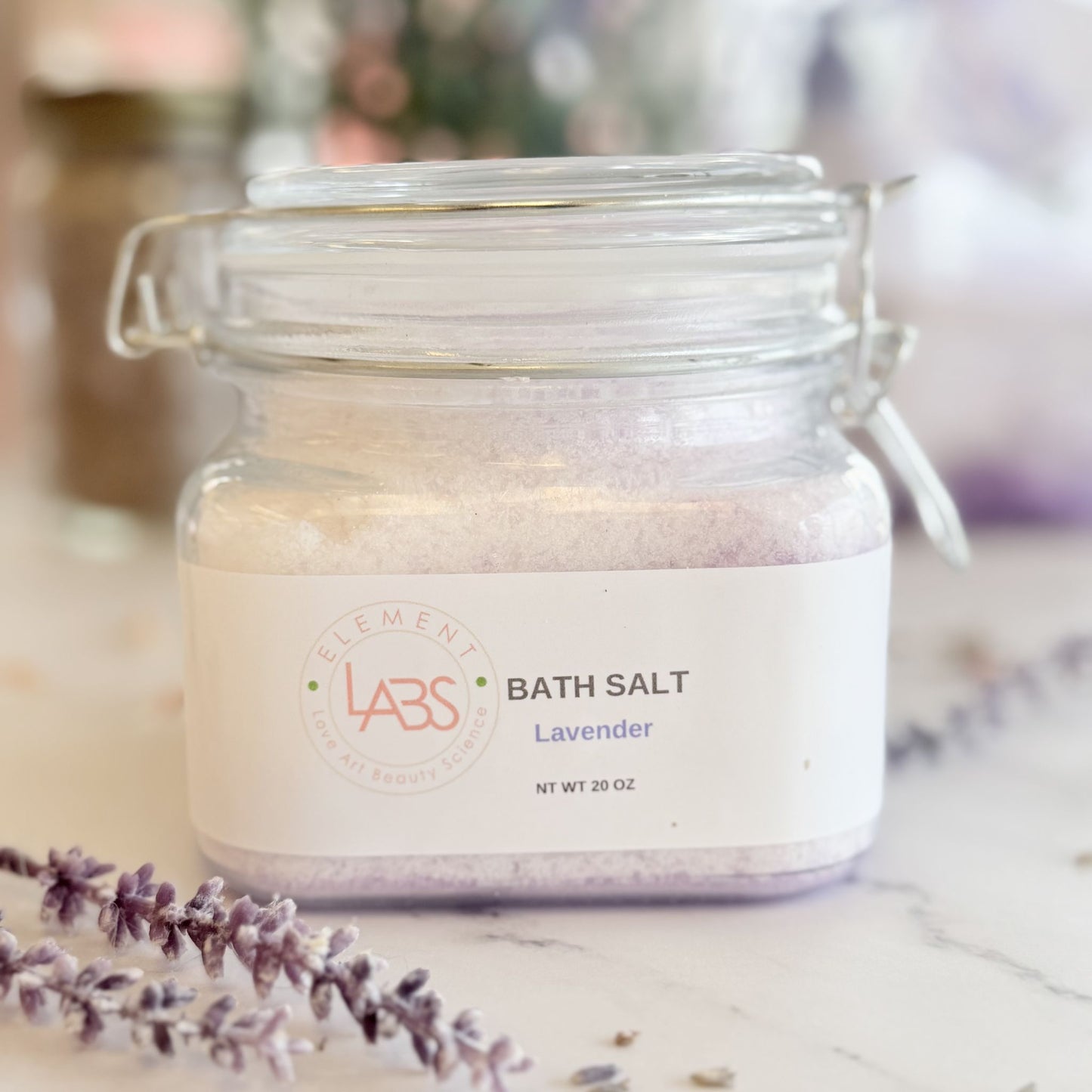 A glass jar of Element Labs Lavender Bath Salt sits on a marble surface with lavender sprigs in the foreground. The clear jar shows light lavender-colored salt inside and has a white label with the brand logo and product name.