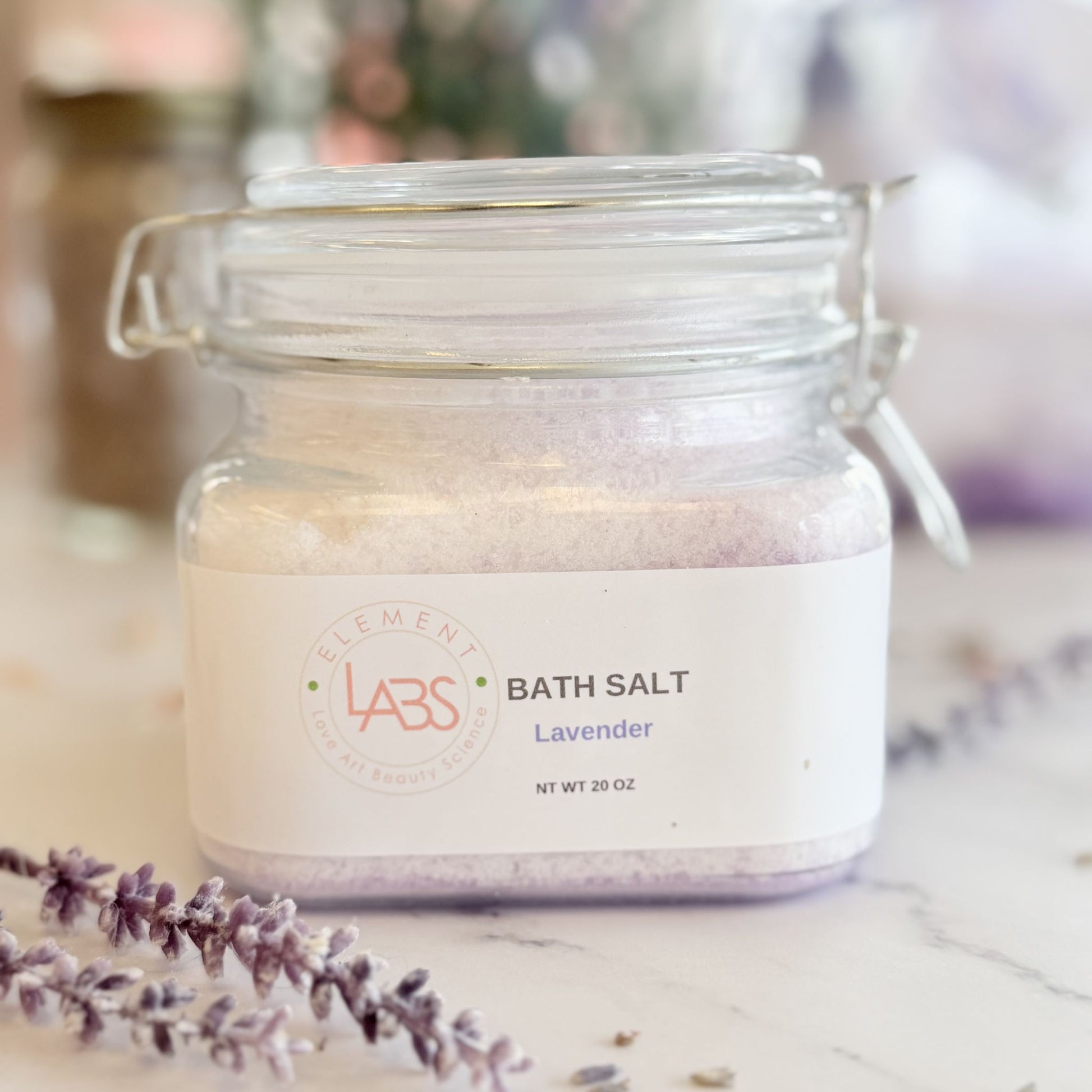 A glass jar of Element Labs Lavender Bath Salt sits on a marble surface with lavender sprigs in the foreground. The clear jar shows light lavender-colored salt inside and has a white label with the brand logo and product name.