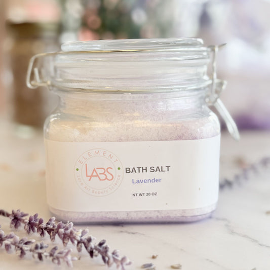 A glass jar of Element Labs Lavender Bath Salt sits on a marble surface with lavender sprigs in the foreground. The clear jar shows light lavender-colored salt inside and has a white label with the brand logo and product name.